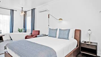 A modern hotel room with a large bed, a desk, a chair, and a bedside table with a lamp.