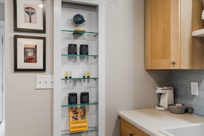 A kitchen with a glass-fronted cabinet displaying kitchenware, a coffee maker, and a framed sign.