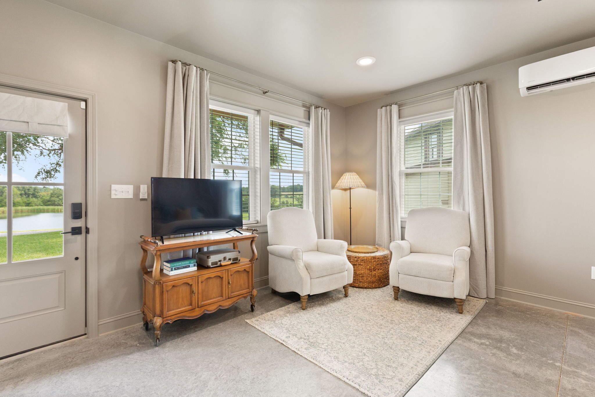 A living room with a flat-screen TV, two armchairs, a small side table, and a large window with curtains.
