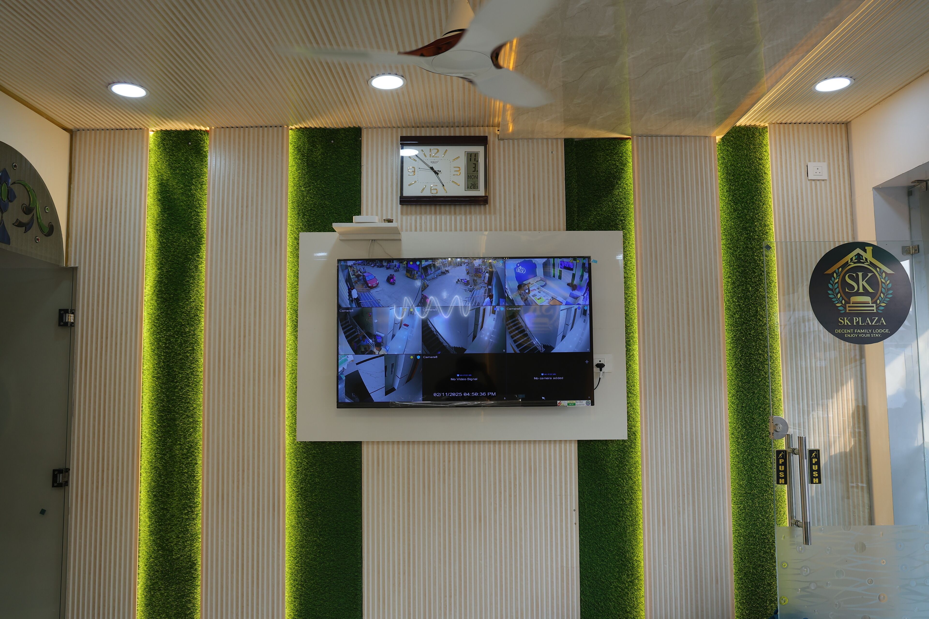 A reception area with a wall-mounted clock, a digital display with multiple camera feeds, and a logo for SK Plaza.