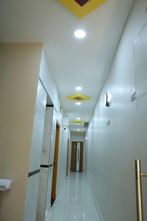 A corridor with a marble wall on one side, a wooden door at the end, and a ceiling with recessed lighting and a yellow accent.