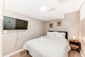 A hotel room with a bed, a bedside table with a lamp, a wall-mounted TV, and a power outlet. - NEW 5BR Home, FREE Private Parking, Entire House (Calgary)