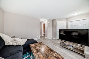 Una sala de estar con un televisor de pantalla plana, un sofá de color oscuro, una mesa de centro y un pasillo que conduce a un baño.