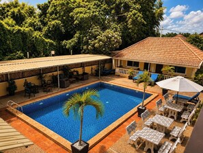 Piscina con sillas de descanso y sombrillas, rodeada por un porche techado y un edificio con techo de tejas.