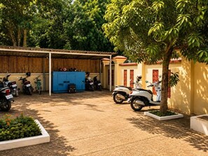 Un área de estacionamiento techada con varias motocicletas aparcadas, un depósito azul y un árbol que da sombra.