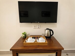 A wooden table with a tray of white cups and saucers, a black kettle, and a small plant. A flat-screen TV is mounted on the wall above the table. - Abode Hotel Siem Reap (Siem Reap)