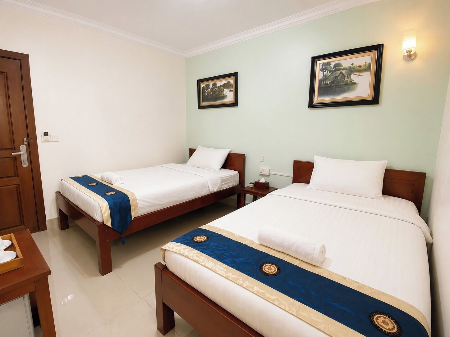 A hotel room with two beds, a wooden door, a small table, and two wall-mounted paintings.