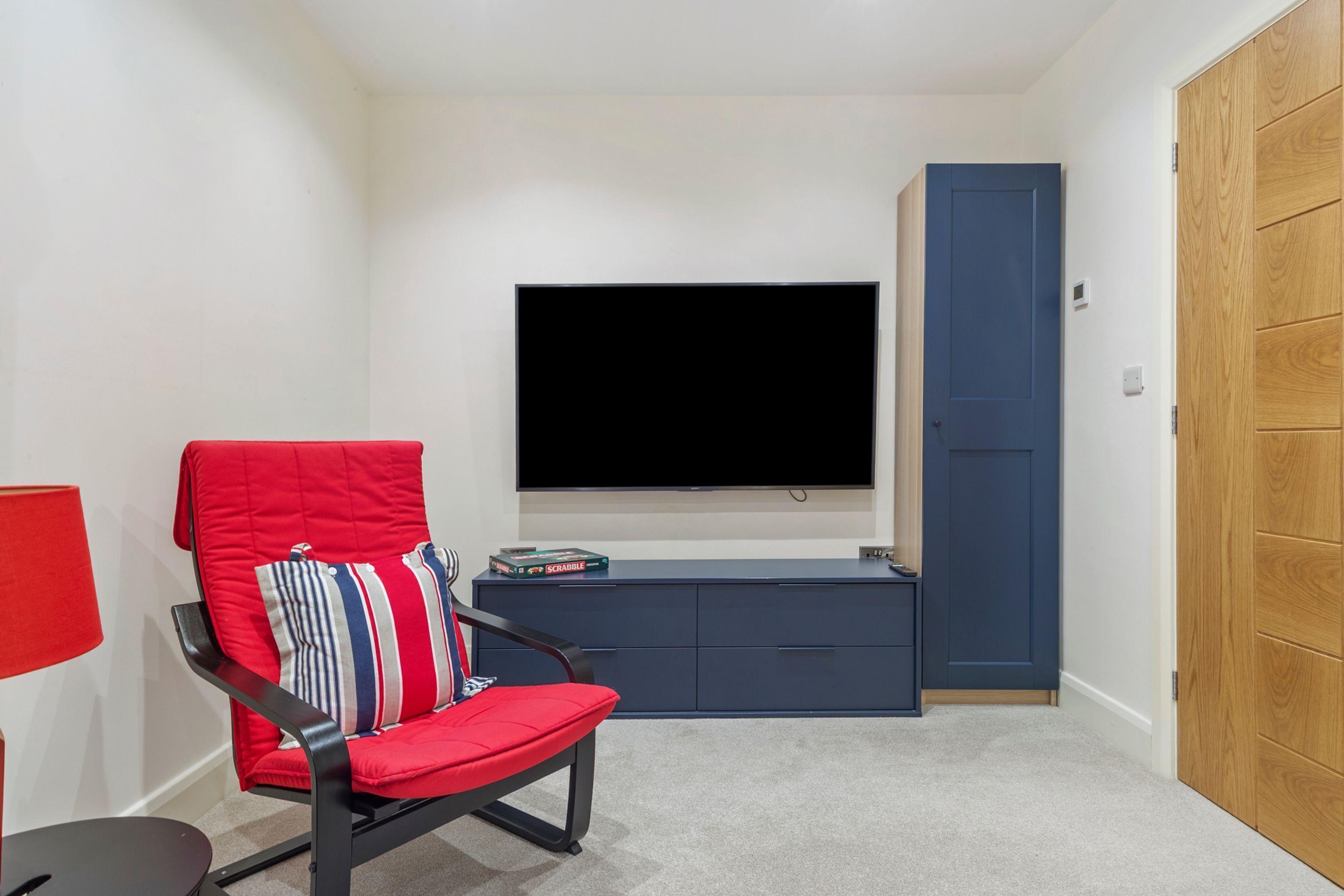 A room with a flat-screen TV mounted on the wall, a red chair with striped cushions, a small blue cabinet, and a wooden door.