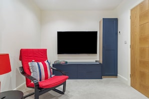 Une pièce avec un téléviseur à écran plat fixé au mur, un fauteuil rouge avec des coussins à rayures, une petite armoire bleue et une porte en bois.