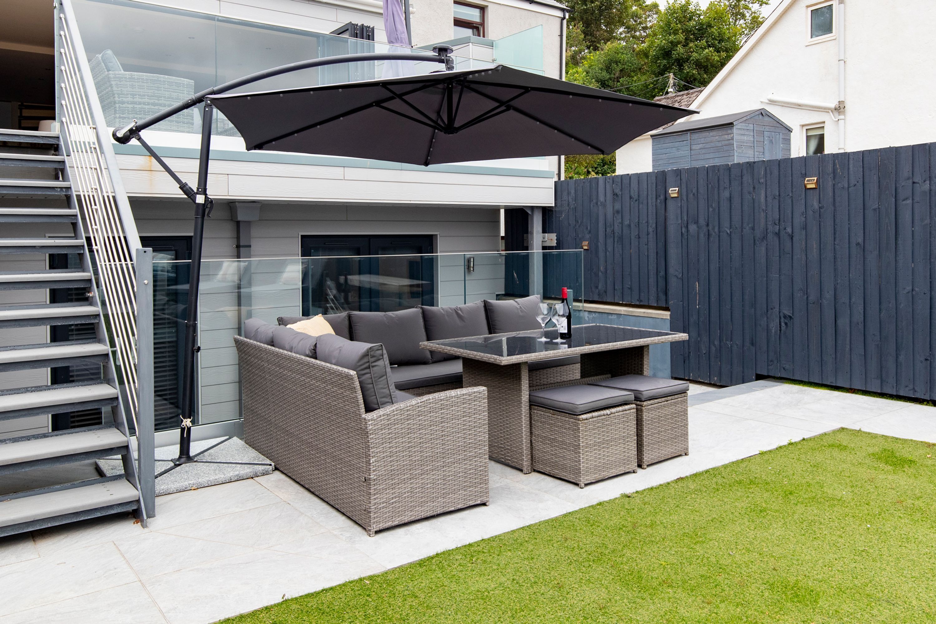 Une terrasse extérieure dotée d'une balustrade en verre, d'un ensemble de meubles de jardin en osier gris, d'une table avec un plateau en verre et d'un parasol foncé.