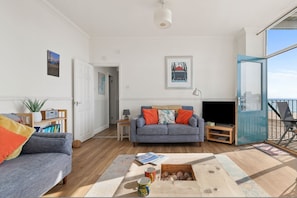 A living room with a grey sofa, a coffee table with books and a mug, a TV, and a view of the sea through a large window. - 1 Sunny Cove - Direct Beach Access, Sea Views (Tenby)