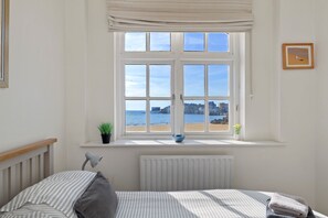A bedroom with a large window, a bed, a radiator, a potted plant, and a framed picture on the wall. - 1 Sunny Cove - Direct Beach Access, Sea Views (Tenby)