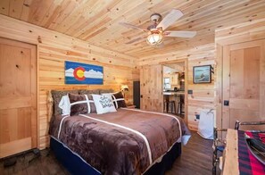 A wooden cabin interior with a bed, a ceiling fan, and a painting of a mountain landscape.