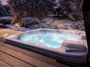Un bain à remous sur une terrasse en bois, entouré d’arbustes et d’arbres couverts de neige.