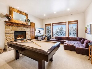 Ein Billardtisch, ein Kamin aus Stein, ein Ecksofa und ein Fernseher in einem Wohnzimmer.