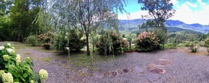A garden with a gravel path and various shrubs, surrounded by trees and a mountain view.