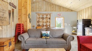 A cozy living room with a sofa, a red chair, a wooden table, and a kitchen area with a refrigerator.