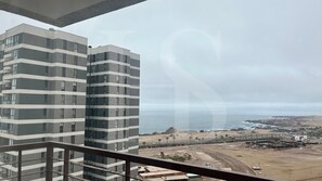 Un edificio alto con múltiples balcones, con vista a una zona costera que incluye una playa y estructuras industriales.