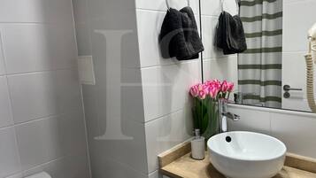 Un baño con paredes de azulejos blancos, un lavabo blanco, un espejo y un perchero con dos toallas.