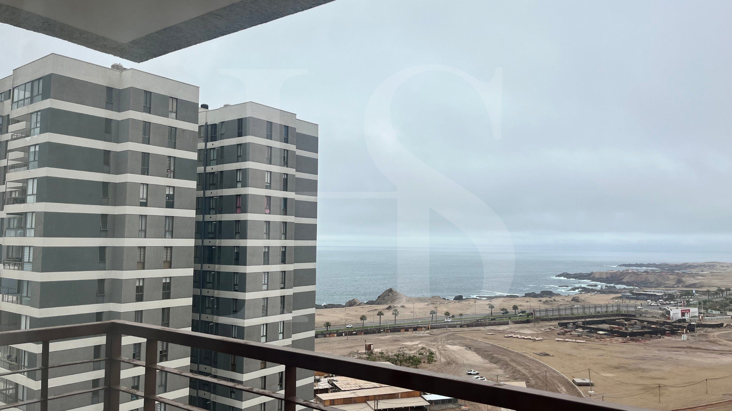 Um edifício alto com várias varandas, com vista para uma área costeira com praia e construções industriais.