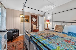A bedroom with a four-poster bed, a desk with a chair, a wooden door, and a nightstand with a lamp.