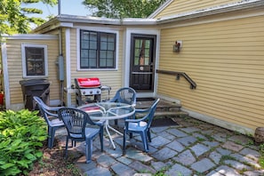 A patio with a glass table and blue chairs, a barbecue grill, and a house with a black door and windows.