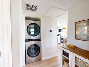 A modern laundry room with a front-loading washer and dryer, a kitchen area in the background, and a wooden console table with a puzzle piece artwork. - Wake up to ocean breezes! (St. Augustine)