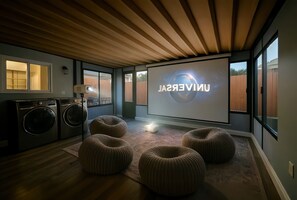 A modern living room with a large screen displaying 'UNIVERSAL,' wooden flooring, a washing machine, and round ottomans. - The Lawndale Cozy Haven (Lawndale)
