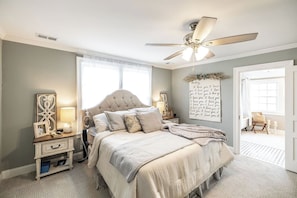 Ein Schlafzimmer mit einer gepolsterten Kopfteil, einem Deckenventilator, einem Nachttisch mit Lampe, einem gerahmten Bild und einer Wand mit einer Dekoration.