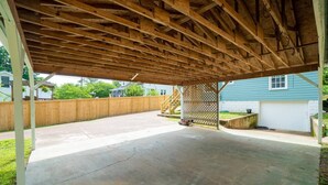 Ett öppet garage med ett träbeklätt tak och ett betonggolv, omgivet av ett trästaket och ett hus i bakgrunden.