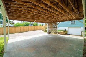 Eine offene Garage mit Holzbalkendecke und Betonboden, umgeben von einem Holzzaun und einem Haus im Hintergrund.