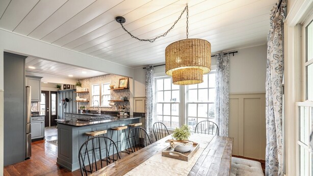 A kitchen with a central island, wooden chairs, a dining table, and a hanging light fixture.