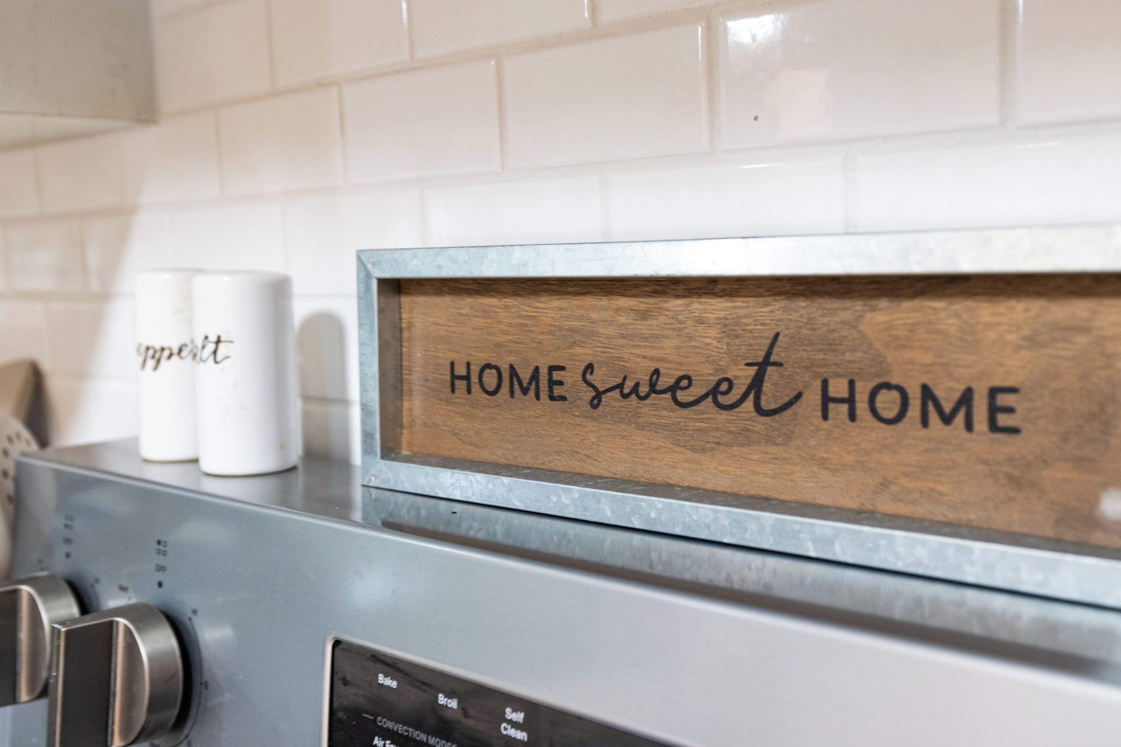 A kitchen with a stainless steel appliance, a wooden sign with 'HOMESWEET HOME' text, and two white containers with text.