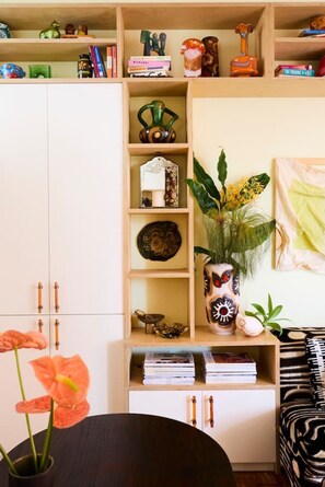 Ein Zimmer mit einem Holzkabinett, Regalen mit Büchern und Dekoration, einer Vase mit Blumen und einem schwarz-weißen Sofabezug.