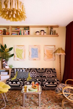 Ein Wohnzimmer mit einem gemusterten Sofa, einem Couchtisch mit Dekoration, einem Bücherregal mit Büchern und dekorativen Objekten sowie einem roten Vorhang.
