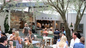 Un espace de restauration en plein air, doté de plusieurs tables et chaises, entouré d’arbres et avec un bâtiment en arrière-plan, dans lequel se trouve une cuisine.
