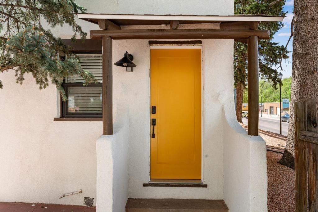Una entrada de edificio con puerta amarilla, una ventana pequeña con persianas, y una estructura de pérgola de madera.