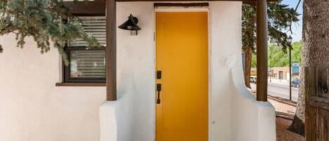 A building entrance with a yellow door, a small window with blinds, and a wooden pergola structure.
