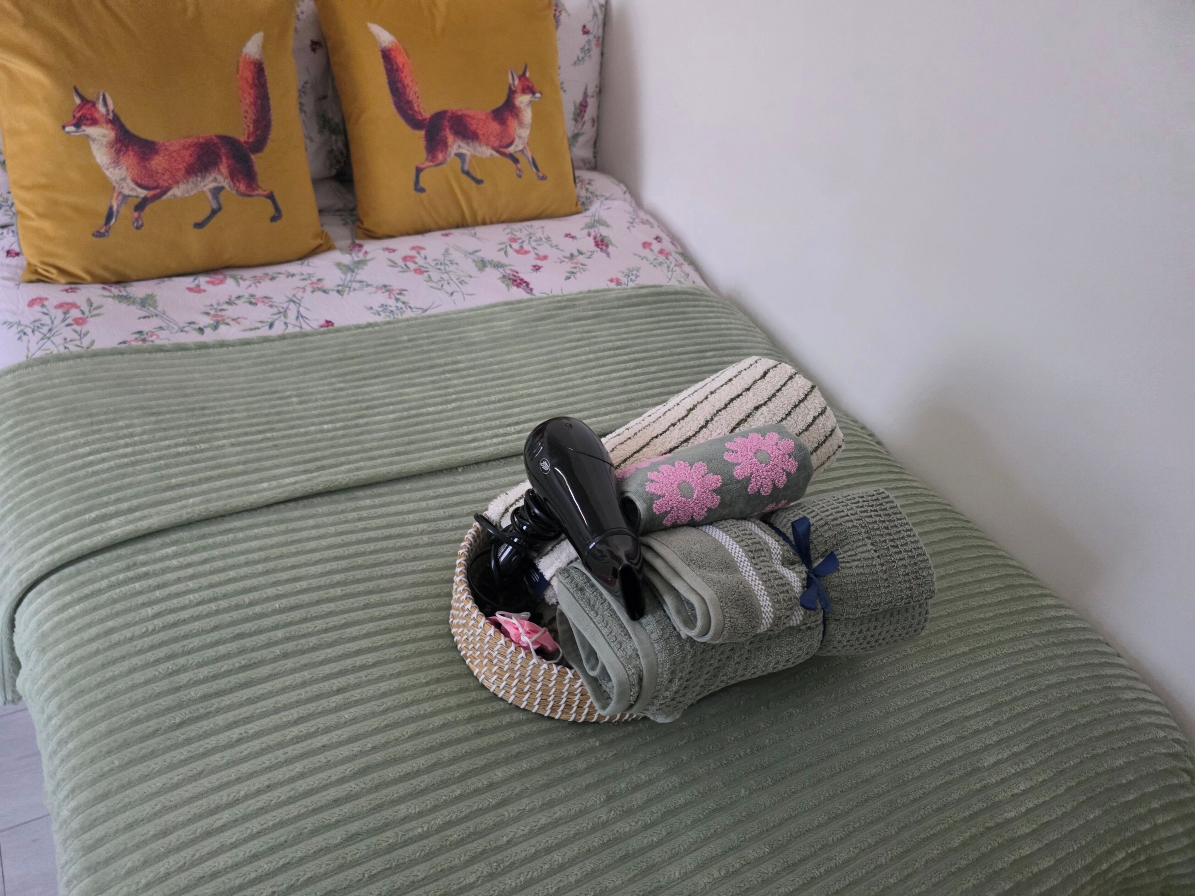 En säng med ett grönt täcke, två kuddar med rävmotiv och ett par skor som ligger i en vävd väska.