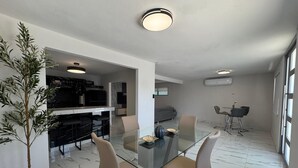 A modern dining area with a glass table and chairs, a kitchen in the background, and a large window providing natural light. - Casa Neeyam: Luxury & space, 7 min from airport. 3BR/2BA, AC, Wi-Fi, parking (Carolina)