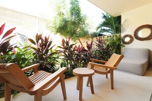 Eine Terrasse mit Holzmöbeln, darunter zwei Stühle und ein kleiner Tisch, umgeben von üppigem Grün und einem grauen Sofa.
