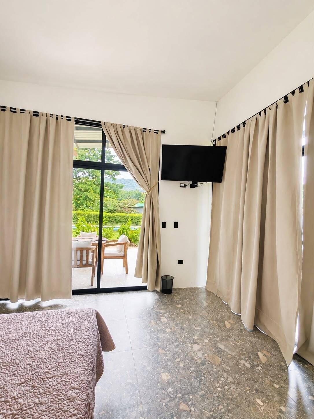 A hotel room with a bed, a television mounted on the wall, and a view of an outdoor area with a table and chairs.