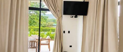 A hotel room with a bed, a television mounted on the wall, and a view of an outdoor area with a table and chairs.