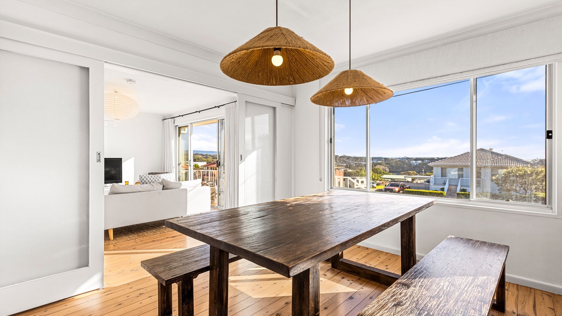 A modern dining area with a wooden table and benches, large windows with a view, and a balcony with a sofa.