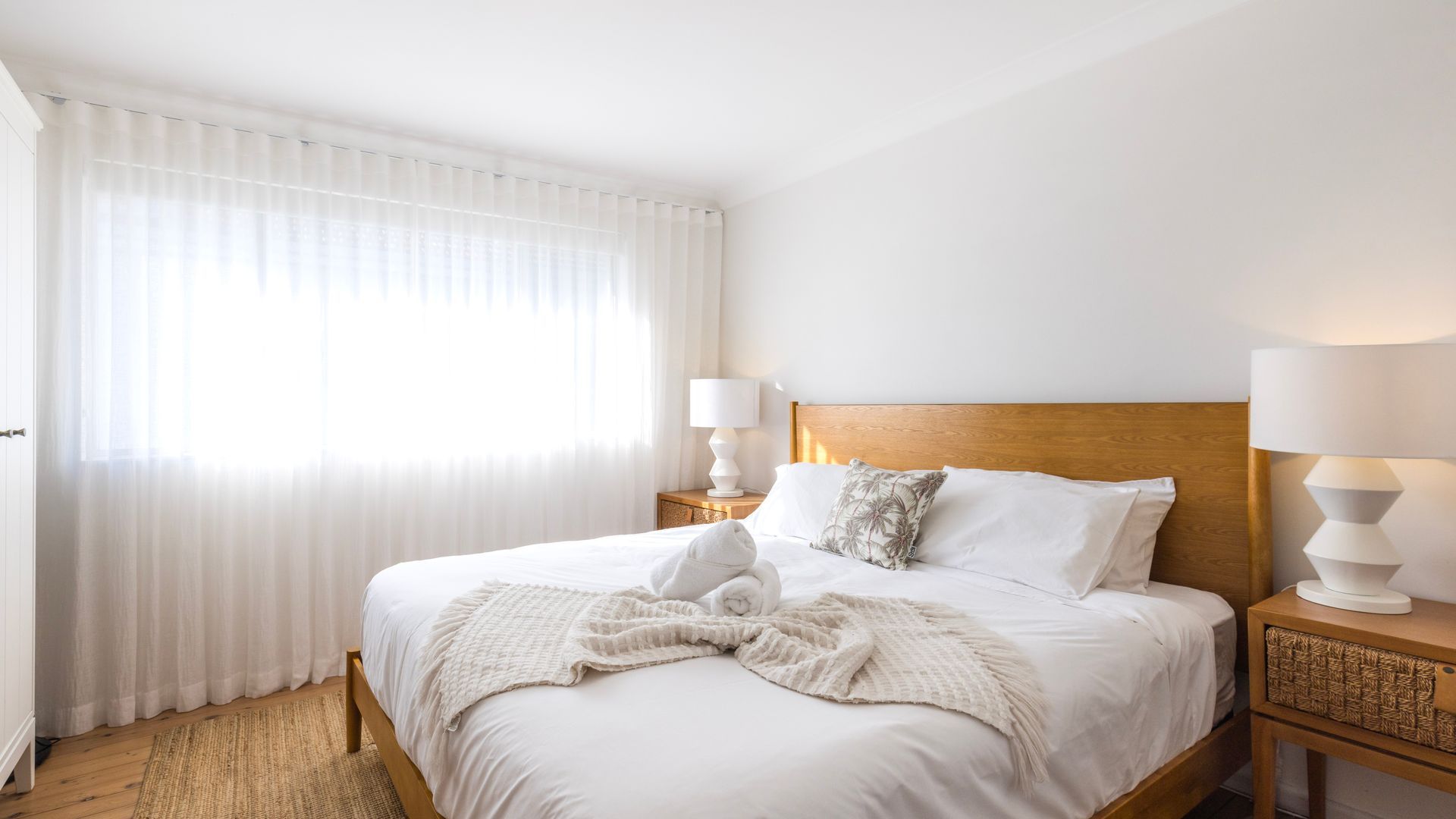 A hotel room with a large bed, two bedside lamps, a wooden headboard, and sheer curtains.