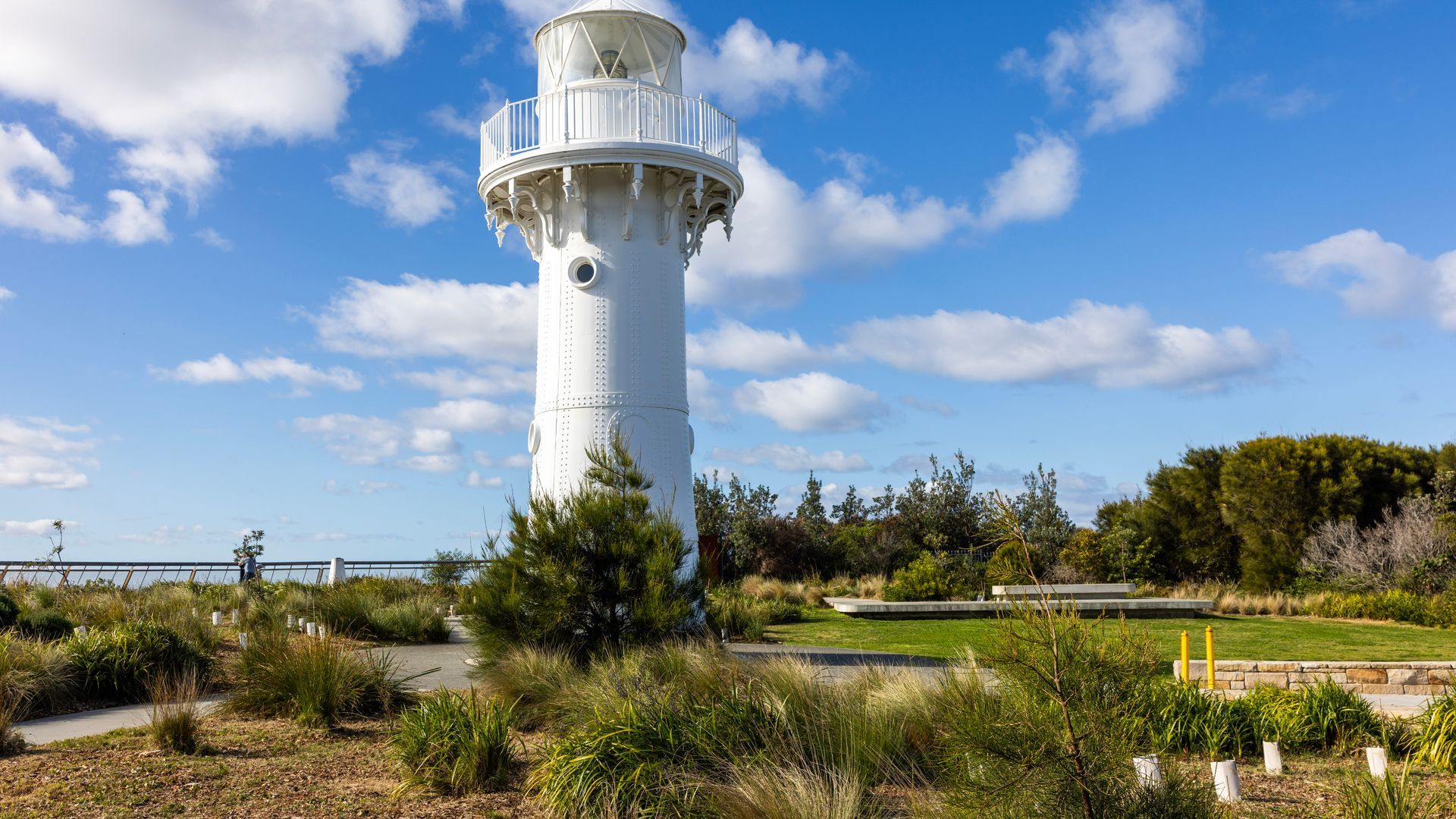 Un phare blanc doté d’une plateforme d’observation circulaire, entouré d’un jardin aménagé avec différents arbustes et d’un sentier menant à celui-ci.