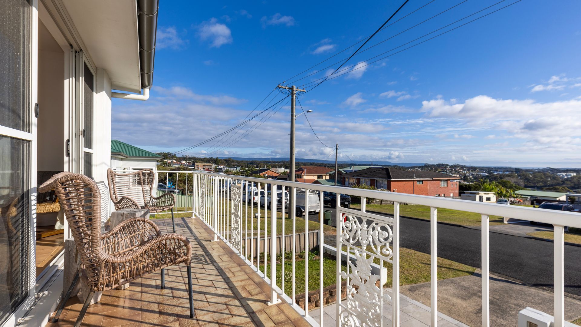 Un balcon avec des fauteuils en osier, une balustrade blanche et une vue sur un quartier résidentiel avec des maisons et une rue.