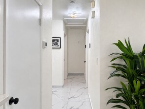 A hallway with white walls, a marble floor, a door with a black handle, a framed picture on the wall, and a potted plant. - 🌴🍹Cozy Retreat 3BR Near Tampa, Disney, Legoland (Lakeland)