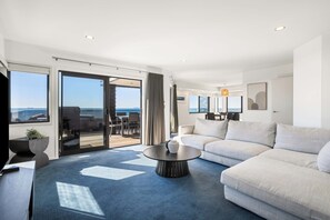 A modern living room with a sofa, a coffee table, and a view of the sea through large windows. - Sunbrae Sands - Mount Maunganui Holiday Homes (Mount Maunganui)