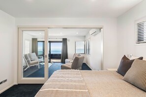 A hotel room with a bed, two chairs, a view of the ocean, and a sliding door leading to a balcony. - Sunbrae Sands - Mount Maunganui Holiday Homes (Mount Maunganui)
